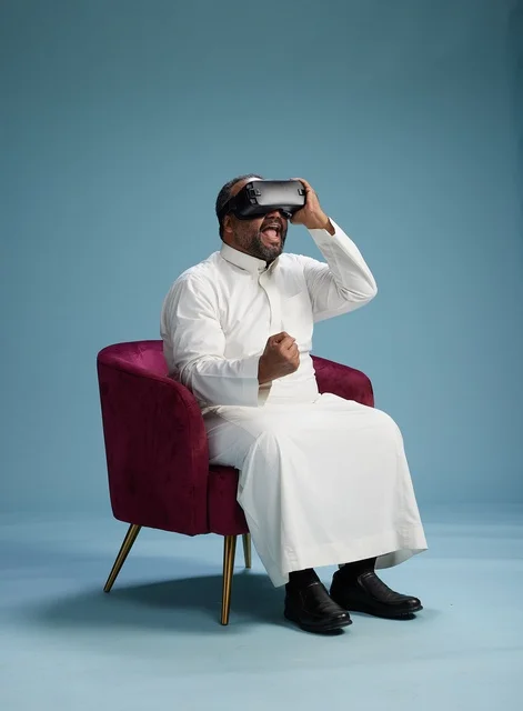 A Saudi man in traditional attire is experiencing virtual reality, watching VR movies, with a blue background.