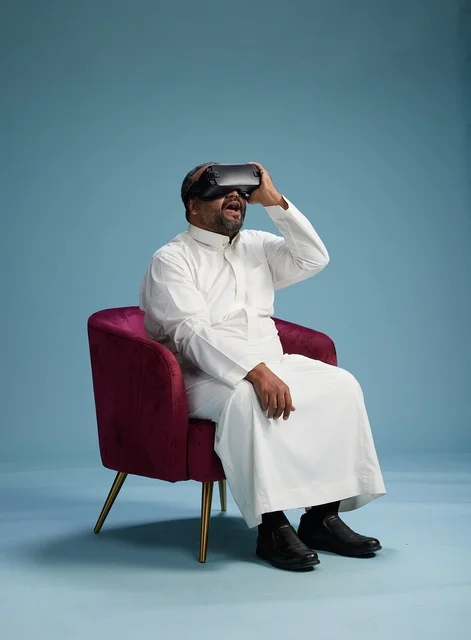 A Saudi man in traditional attire is experiencing virtual reality, watching VR movies, with a blue background.