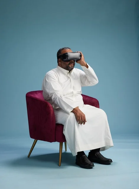 A Saudi man in traditional attire is experiencing virtual reality, watching VR movies, with a blue background.