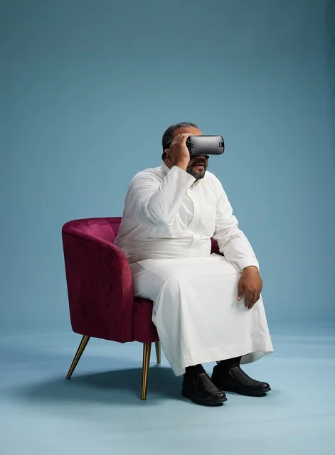 A Saudi man in traditional attire is experiencing virtual reality, watching VR movies, with a blue background.