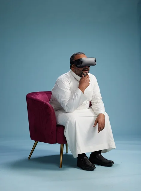 A Saudi man in traditional attire is experiencing virtual reality, watching VR movies, with a blue background.