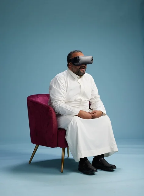 A Saudi man in traditional attire is experiencing virtual reality, watching VR movies, with a blue background.