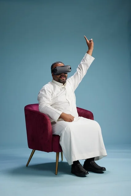 A Saudi man in traditional attire is experiencing virtual reality, watching VR movies, with a blue background.