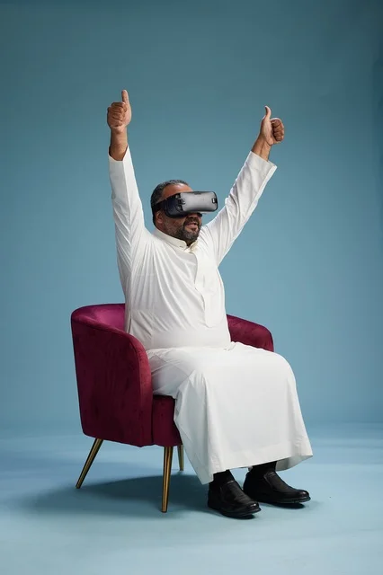 A Saudi man in traditional attire is experiencing virtual reality, watching VR movies, with a blue background.