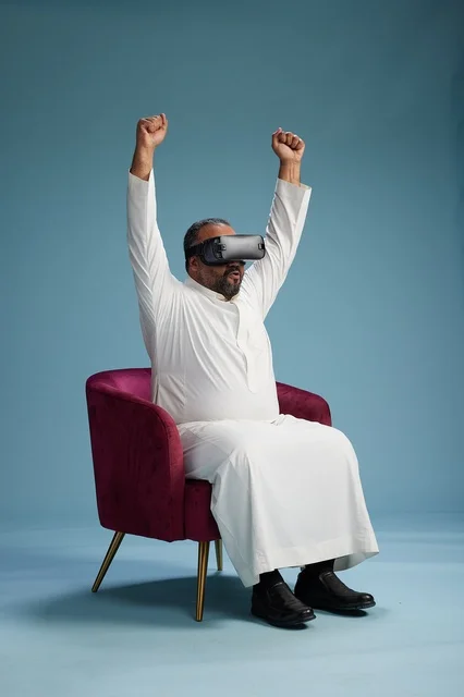 A Saudi man in traditional attire is experiencing virtual reality, watching VR movies, with a blue background.