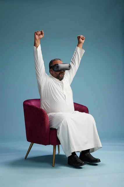 A Saudi man in traditional attire is experiencing virtual reality, watching VR movies, with a blue background.