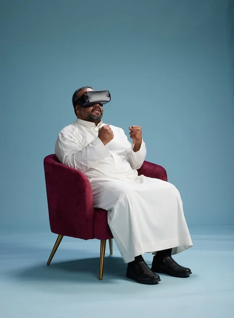 A Saudi man in traditional attire is experiencing virtual reality, watching VR movies, with a blue background.