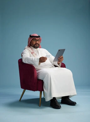 رجل  عربي سعودي خليجي يرتدي الثوب الابيض والشماغ  يتسوق عبر الإنترنت باستخدام جهاز لوحي وبطاقة ائتمان, الجلوس على كرسي احمر, خلفيه زرقاء