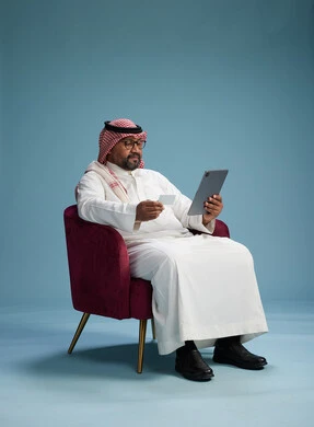 رجل  عربي سعودي خليجي يرتدي الثوب الابيض والشماغ  يتسوق عبر الإنترنت باستخدام جهاز لوحي وبطاقة ائتمان, الجلوس على كرسي احمر, خلفيه زرقاء