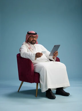 A Saudi Arabian Gulf Arab man wearing a white thobe and a shemagh is shopping online using a tablet and a credit card, sitting on a red chair, with a blue background.