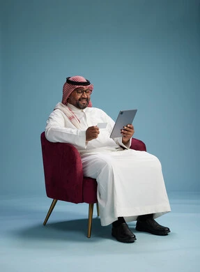 A Saudi Arabian Gulf Arab man wearing a white thobe and a shemagh is shopping online using a tablet and a credit card, sitting on a red chair, with a blue background.