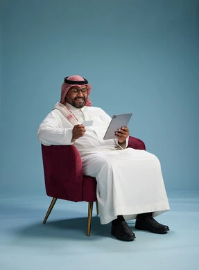A Saudi Arabian Gulf Arab man wearing a white thobe and a shemagh is shopping online using a tablet and a credit card, sitting on a red chair, with a blue background.
