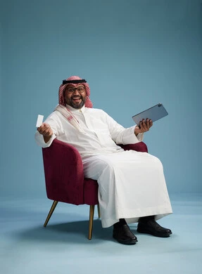 رجل  عربي سعودي خليجي يرتدي الثوب الابيض والشماغ  يتسوق عبر الإنترنت باستخدام جهاز لوحي وبطاقة ائتمان, الجلوس على كرسي احمر, خلفيه زرقاء