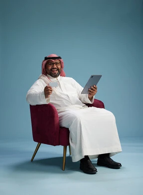 رجل  عربي سعودي خليجي يرتدي الثوب الابيض والشماغ  يتسوق عبر الإنترنت باستخدام جهاز لوحي وبطاقة ائتمان, الجلوس على كرسي احمر, خلفيه زرقاء