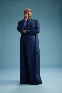 A portrait of a Saudi man standing wearing a navy blue thobe, making various gestures and movements, standing full body, with a blue background.