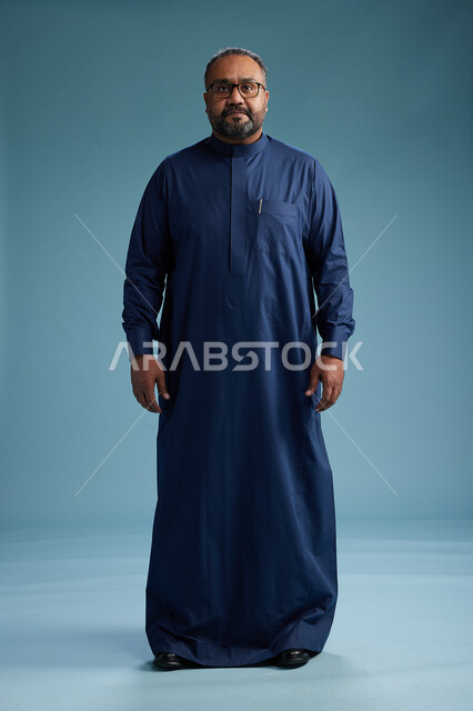 بورتريه لرجل سعودي يقف مرتدياً ثوب كحلي يقوم بايماءات وحركات متنوعه, الوقوف بكامل الجسم, خلفيه زرقاء