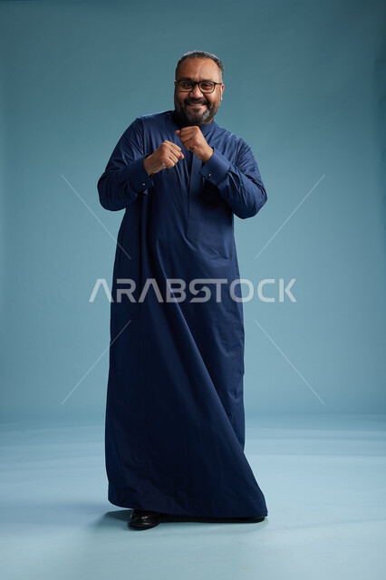 بورتريه لرجل سعودي يقف مرتدياً ثوب كحلي يقوم بايماءات وحركات متنوعه, الوقوف بكامل الجسم, خلفيه زرقاء