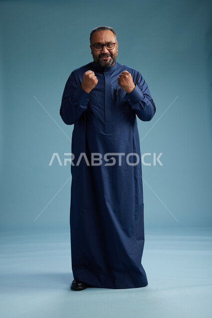 بورتريه لرجل سعودي يقف مرتدياً ثوب كحلي يقوم بايماءات وحركات متنوعه, الوقوف بكامل الجسم, خلفيه زرقاء