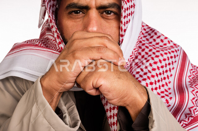 بورتريه لرجل عربي خليجي سعودي يرتدي ملابس الشتاء الدافئة وينفخ بيده، الشعور بالصقيع، ارتداء جاكيت الشتاء، التنفس على اليدين لابقائها دافئة، ايماءات وجه ويدين تدل على البرد الشديد، اجواء شتوية، خلفية بيضاء