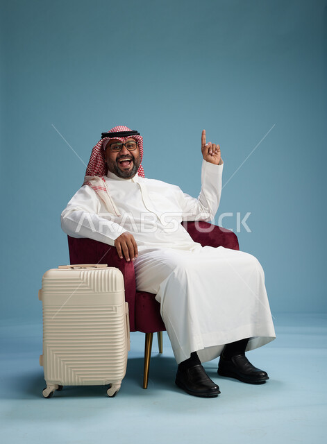 بورتريه لرجل سعودي  يرتدي الثوب الابيض والشماغ يجلس مع حقيبة سفر بيضاء, مفهوم السياحه والسفر, خلفيه زرقاء