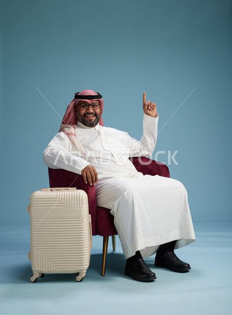 بورتريه لرجل سعودي  يرتدي الثوب الابيض والشماغ يجلس مع حقيبة سفر بيضاء, مفهوم السياحه والسفر, خلفيه زرقاء