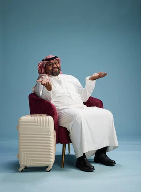 A portrait of a Saudi man wearing a white thobe and a shemagh sitting with a white travel bag, the concept of tourism and travel, blue background.