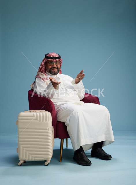 بورتريه لرجل سعودي  يرتدي الثوب الابيض والشماغ يجلس مع حقيبة سفر بيضاء, مفهوم السياحه والسفر, خلفيه زرقاء
