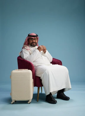A portrait of a Saudi man wearing a white thobe and a shemagh sitting with a white travel bag, the concept of tourism and travel, blue background.