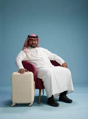 A portrait of a Saudi man wearing a white thobe and a shemagh sitting with a white travel bag, the concept of tourism and travel, blue background.