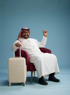 A portrait of a Saudi man wearing a white thobe and a shemagh sitting with a white travel bag, the concept of tourism and travel, blue background.
