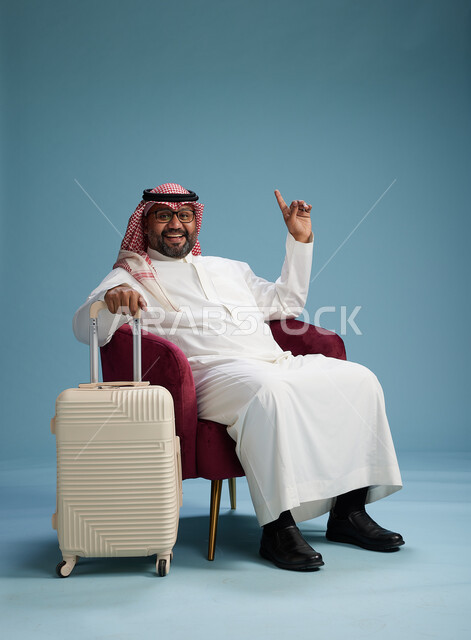 A portrait of a Saudi man wearing a white thobe and a shemagh sitting with a white travel bag, the concept of tourism and travel, blue background.