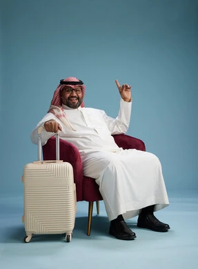 A portrait of a Saudi man wearing a white thobe and a shemagh sitting with a white travel bag, the concept of tourism and travel, blue background.