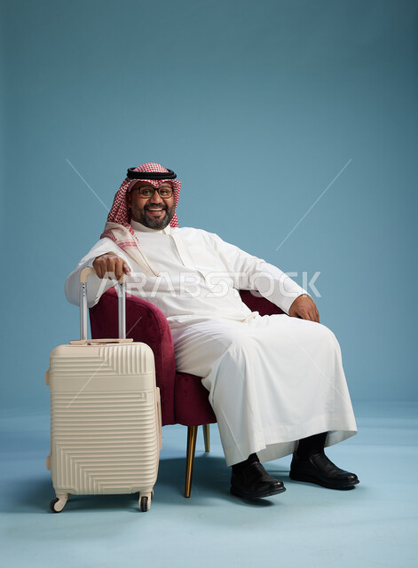 بورتريه لرجل سعودي  يرتدي الثوب الابيض والشماغ يجلس مع حقيبة سفر بيضاء, مفهوم السياحه والسفر, خلفيه زرقاء