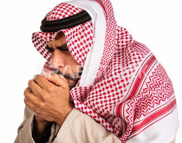 بورتريه لرجل عربي خليجي سعودي يرتدي ملابس الشتاء الدافئة وينفخ بيده، الشعور بالصقيع، ارتداء جاكيت الشتاء، التنفس على اليدين لابقائها دافئة، ايماءات وجه ويدين تدل على البرد الشديد، اجواء شتوية، خلفية بيضاء
