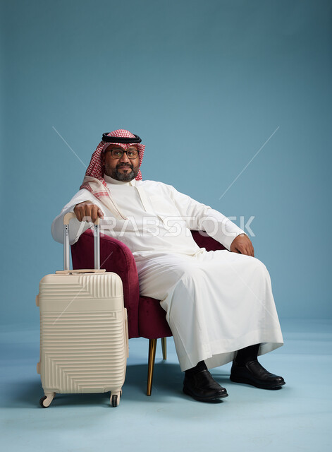 A portrait of a Saudi man wearing a white thobe and a shemagh sitting with a white travel bag, the concept of tourism and travel, blue background.