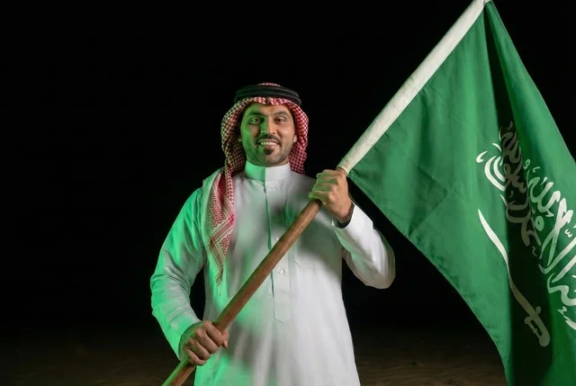 إحياء ذكرى اليوم الوطني السعودي 23 سبتمبر، يوم العلم السعودي 11 مارس, إيماءات الفخر و السرور بالانتماء للوطن, رجل عربي سعودي يرتدي الشماغ والثوب التقليدي يحمل بيده علم المملكة، يوم بدينا 1727 م