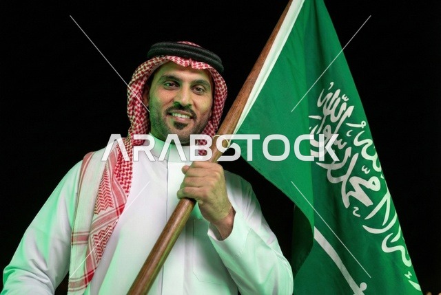 Commemoration of the Saudi National Day on September 23, Saudi Flag Day on March 11, gestures of pride and joy in belonging to the homeland, an Arab Saudi man wearing the traditional ghutrah and thobe holding the flag of the Kingdom in his hand, the day we began in 1727 AD.