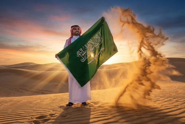 يوم العلم السعودي 11 مارس, إحياء ذكرى اليوم الوطني السعودي 23 سبتمبر، إيماءات الفخر و السرور بالانتماء للوطن, رجل عربي سعودي يرتدي الشماغ والثوب التقليدي يحمل بيده علم المملكة، يوم بدينا 1727 م ، خلفية شروق الشمس في الصحراء