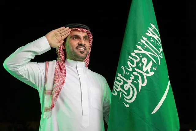 Gestures of pride and joy in belonging to the homeland, commemorating Saudi National Day on September 23, Saudi Flag Day on March 11, an Arab Saudi man wearing the ghutrah and traditional thobe greeting in front of the Kingdom's flag, the day we began in 1727 AD, with a background of sunrise in the desert.