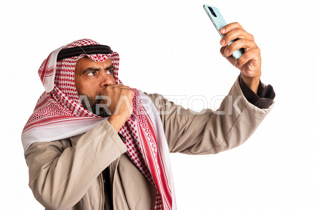 بورتريه لرجل عربي خليجي سعودي يرتدي ملابس الشتاء الدافئة، التقاط صورة تذكارية من خلال الهاتف المحمول، صور تذكارية، صور سيلفي، كاميرا امامية، مكالمة فيديو، بث فيديو مباشر، ايماءات وجه ويدين تدل على البرد الشديد، اجواء شتوية، خلفية بيضاء