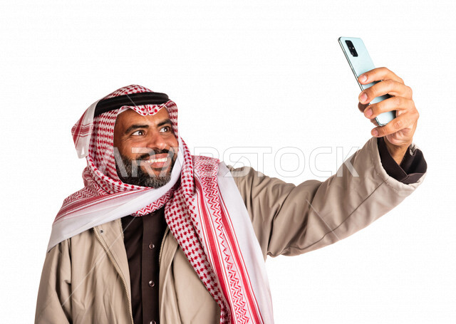 بورتريه لرجل عربي خليجي سعودي يرتدي ملابس الشتاء الدافئة، التقاط صورة تذكارية من خلال الهاتف المحمول، صور تذكارية، صور سيلفي، كاميرا امامية، ايماءات وجه مبتسمة، مكالمة فيديو ، بث فيديو مباشر، اجواء شتوية، خلفية بيضاء