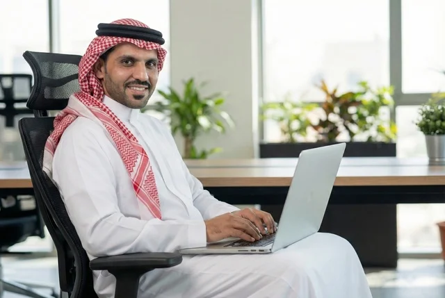Completing the required tasks, Saudi office professions and jobs, managing and organizing company affairs through technical devices, integrating work with technology, an Arab Gulf Saudi businessman wearing traditional attire and a ghutrah working on a laptop, working in the economic field.
