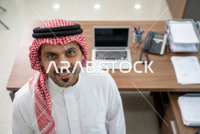 Completing the required tasks, Saudi office professions and jobs, managing and organizing company affairs through technical devices, integrating work with technology, an Arab Gulf Saudi businessman wearing traditional attire and a ghutrah looking at the camera with gestures of happiness, working on a laptop, working in the economic field.