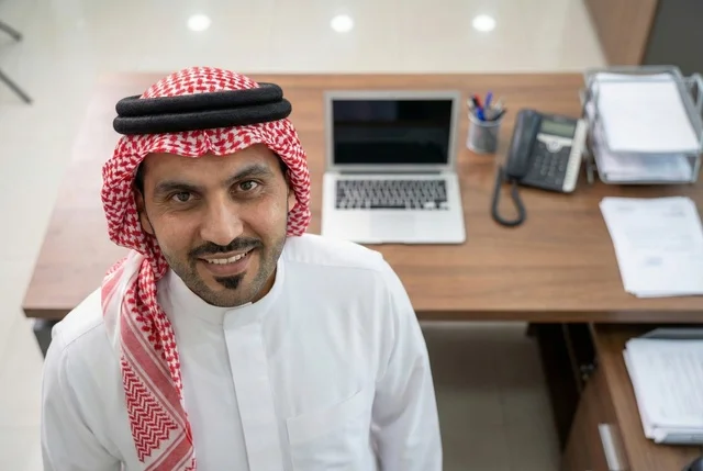 Completing the required tasks, Saudi office professions and jobs, managing and organizing company affairs through technical devices, integrating work with technology, an Arab Gulf Saudi businessman wearing traditional attire and a ghutrah looking at the camera with gestures of happiness, working on a laptop, working in the economic field.