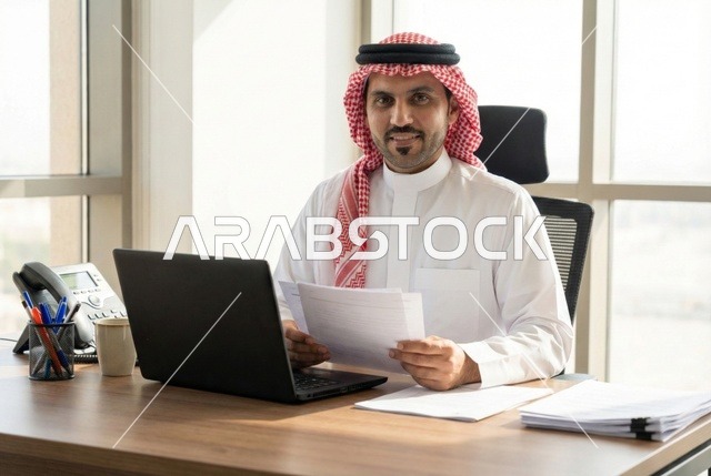 Integrating technology and modern techniques in the office and administrative sector, managing business via a laptop, supervising work, a portrait of an Arab Gulf Saudi man wearing traditional attire and a ghutrah sitting at a modern desk with a set of plans and papers in front of him.
