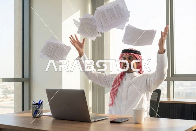 Integrating technology and modern techniques in the office and administrative sector, managing business via a laptop, supervising work, a portrait of an Arab Gulf Saudi man wearing traditional attire and a ghutrah sitting at a modern desk with a set of plans and papers in front of him.