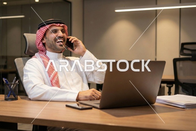 اجراء مكالمة هاتفية، دمج التكنولوجيا والتقنيات الحديثة في القطاع المكتبي والإداري، إدارة الأعمال عبر الحاسوب المحمول، الإشراف على العمل، بورتريه  لرجل عربي خليجي سعودي يرتدي الثوب التقليدي والشماغ يجلس على مكتب عصري وأمامه مجموعة مخططات واوراق