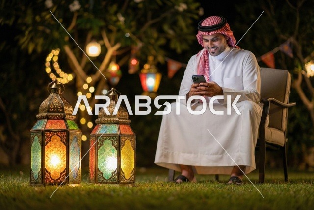 Using a mobile device, an Arab Gulf Saudi man wearing traditional Saudi attire is using a mobile phone while sitting outdoors, engaging in social communication and browsing websites, Ramadan lantern, lighting and decorations of the month of Ramadan, Ramadan atmosphere.