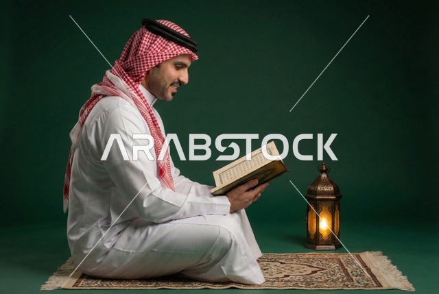 رجل عربي خليجي سعودي  يرتدي الزي التقليدي والشماغ يقرأ القرآن الكريم، المصحف الشريف، تلاوة وحفظ القران الكريم، فانوس رمضان، استقبال شهر رمضان المبارك، زينة شهر رمضان، أجواء رمضانية، جلسة خارجية في الهواء الطلق