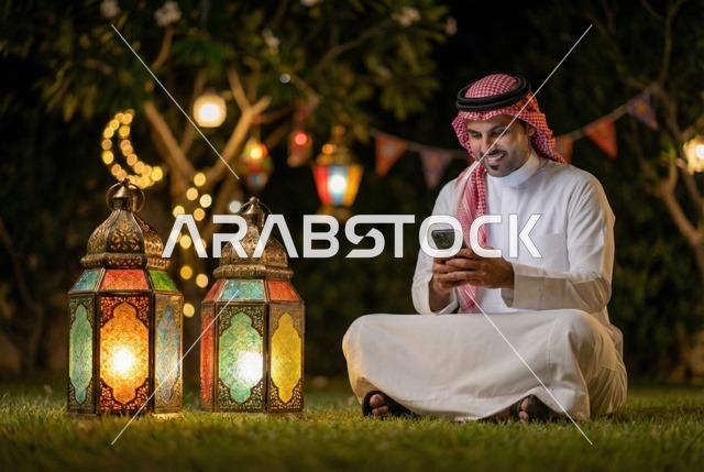 استخدام جهاز الموبايل ، رجل عربي خليجي سعودي يرتدي الزي السعودي التقليدي يستخدم الهاتف المحمول يجلس في الهواء الطلق في الخارج ، التواصل الاجتماعي وتصفح المواقع الالكترونية، فانوس رمضان، إضاءة وزينة شهر رمضان، أجواء رمضانية